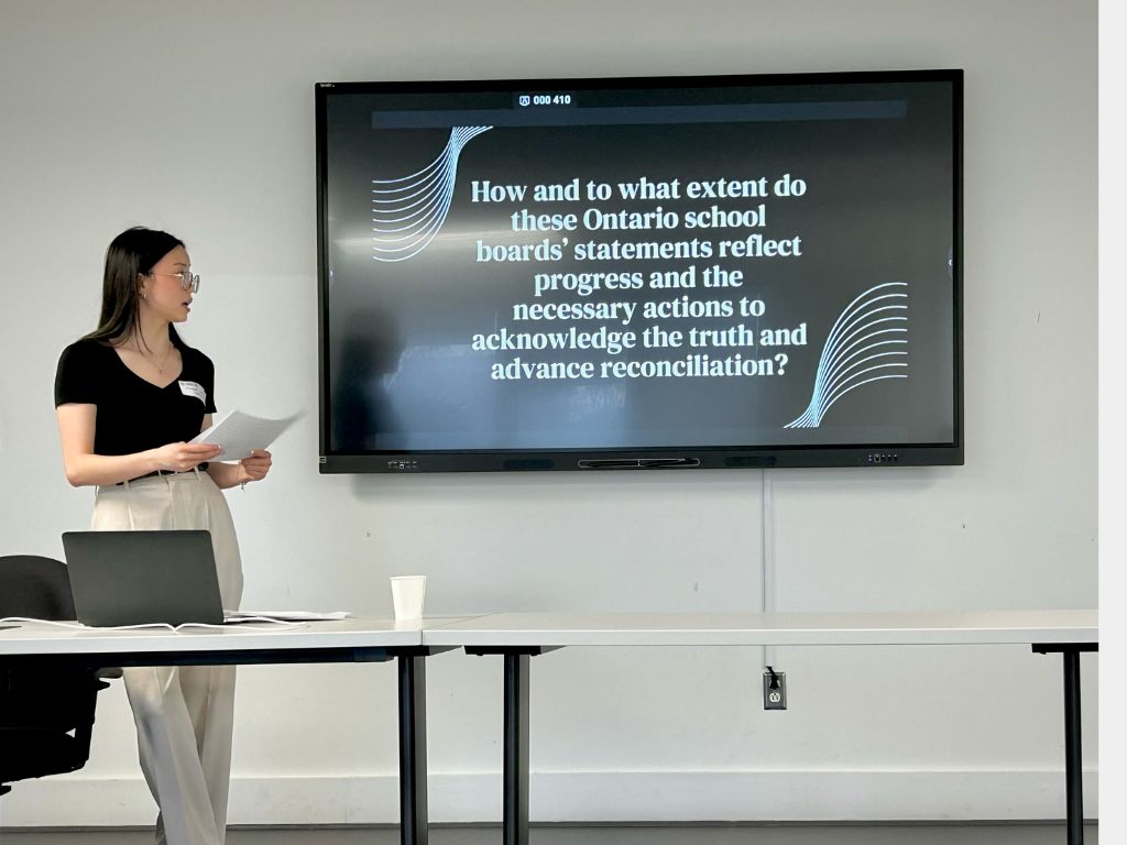 Student standing in front of presentation slide that reads: “How and to what extent do these Ontario school boards’ statements reflect progress and the necessary actions to acknowledge the truth and advance reconciliation?”